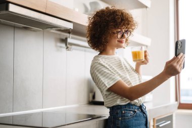 Gülümseyen kıvırcık kadın ev mutfağında cep telefonuyla selfie çekerken meyve suyu içiyor.
