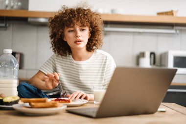 Avrupalı genç bir kadın evde kahvaltı yaparken dizüstü bilgisayar kullanıyor.
