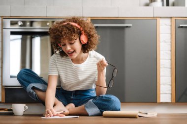 Mutfakta yerde otururken kulaklıklı, gülümseyen kıvırcık kadın resim çiziyor.