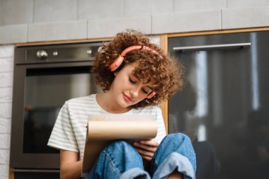 Mutfakta yerde otururken kulaklıklı, gülümseyen kıvırcık kadın resim çiziyor.