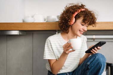 Mutfakta kahve içerken kulaklıkla gülümseyen kıvırcık kadın.