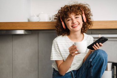Mutfakta kahve içerken kulaklıkla gülümseyen kıvırcık kadın.