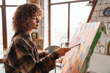 Stüdyo atölyesinde tuvalin önünde duran gülümseyen kızıl saçlı kadın ressam.