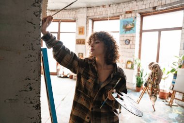 Stüdyo atölyesinde tuvalin önünde duran gülümseyen kızıl saçlı kadın ressam.