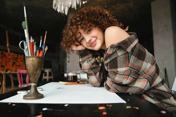 若い赤毛の女性アーティストを笑顔で描き 鉛筆で机の上に座ってお茶を飲みながらスタジオワークショップ — ストック写真