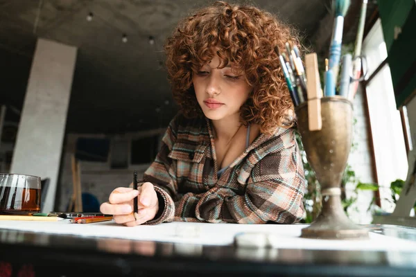 若い赤毛の女性アーティストは スタジオのワークショップでお茶のカップと机に座って鉛筆で描く — ストック写真