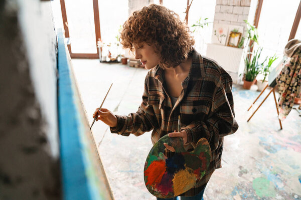 Smiling young white redhead woman artist painting standing in front of canvas in studio workshop