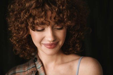 Gülümseyen kızıl saçlı, ekose gömlekli bir kadın stüdyoda dikilip poz veriyor.