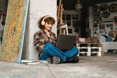 Bir sanat stüdyosunda dizüstü bilgisayarla oturan, kulaklıklı, gülümseyen, kızıl saçlı bir kadın.