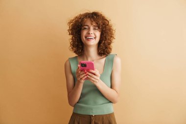 Bej arka planda izole edilmiş cep telefonuyla gülen genç bir kızıl kadın.