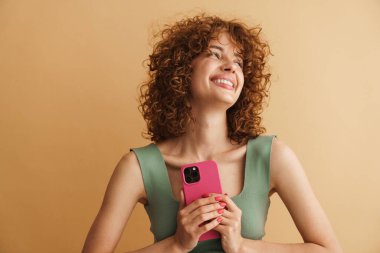 Bej arka planda izole edilmiş cep telefonuyla gülen genç bir kızıl kadın.