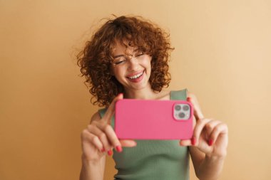 Bej arka planda izole edilmiş cep telefonuyla fotoğraf çekerken gülümseyen genç kızıl kadın.