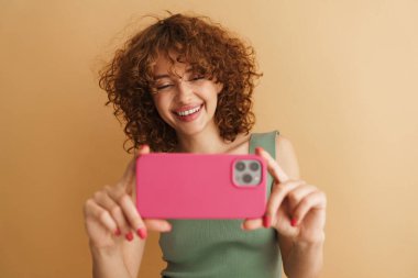 Bej arka planda izole edilmiş cep telefonuyla fotoğraf çekerken gülümseyen genç kızıl kadın.