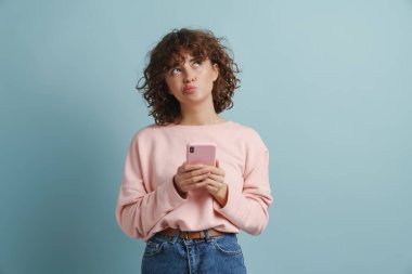 Genç kızıl, kafası karışmış bir kadın cep telefonu kullanıyor ve mavi arka planda tek başına yukarıya bakıyor.