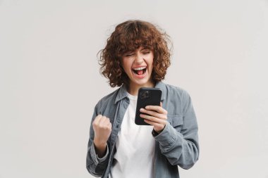 Heyecanlı genç bir kadın, beyaz arka planda izole edilmiş cep telefonu kullanırken kazanan hareketi yapıyor.