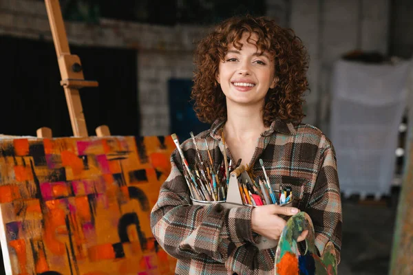 Sonriente Joven Pelirroja Blanca Pintando Pie Delante Lienzo Sosteniendo Pinceles — Foto de Stock