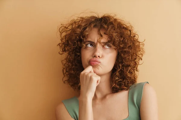 Young ginger curly woman frowning while looking upward isolated over beige background