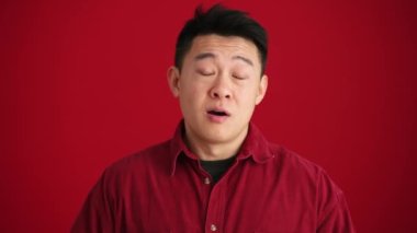 Tişört giyen neşeli Asyalı adam bir şeye katılıyor ve kırmızı stüdyoda el hareketi yapıyor.