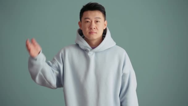 Handsome Asian Man Wearing Hoodie Waving Camera Blue Studio — Stock Video