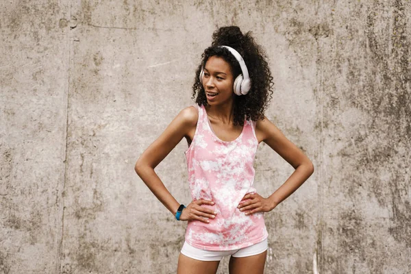 年轻的黑人女运动员站在混凝土墙边用耳机听音乐 — 图库照片