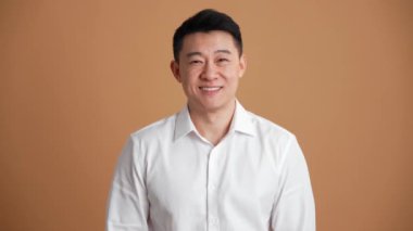 Beyaz gömlek giyen neşeli Asyalı adam turuncu stüdyodaki kameraya bakıyor.