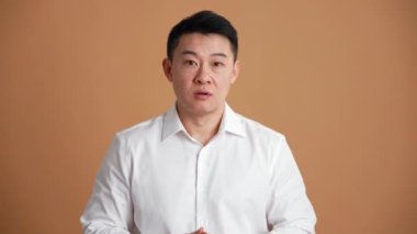 Beyaz tişört giyen mutlu Asyalı bir adam bir şeye katılıyor ve turuncu stüdyoda el hareketi yapıyor.