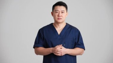 Üzgün Asyalı doktor gri stüdyoda kameraya konuşuyor.