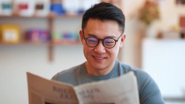 陽気なアジアの若者が眼鏡をかけてカフェで新聞を読んでいます — ストック動画