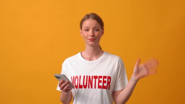 Smiling Woman Volunteer Waving Camera Pointing Phone Orange Studio — Stok Video