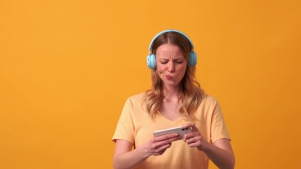 Blondýnka Nosí Žluté Tričko Hraje Hru Telefonu Sluchátkách Rozčiluje Oranžovém — Stock video