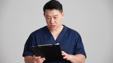Güzel gülümseyen Asyalı doktor hastalıkların tarihini okuyor ve gri stüdyoda kameraya konuşuyor.