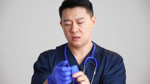 Κουρασμένος Ασιάτης Γιατρός Αναβάλλει Γάντια Στο Γκρίζο Στούντιο — Αρχείο Βίντεο