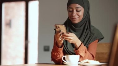 Olumlu Müslüman bir kadın evdeki masada bir fincan çayla telefonla mesaj atıyor.