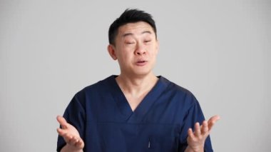 Yakışıklı gülümseyen Asyalı doktor gri stüdyoda kameraya konuşuyor.