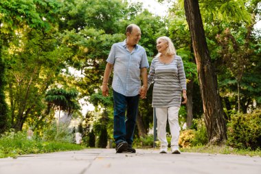 Yaz parkında yürürken el ele tutuşan ve gülümseyen beyaz yaşlı çift.