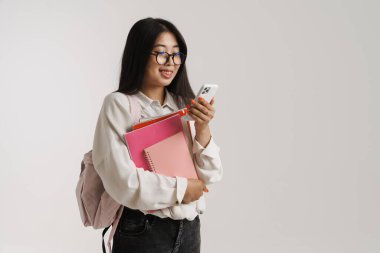 Genç Asyalı kadın beyaz duvarda izole edilmiş egzersiz kitaplarıyla poz verirken cep telefonu kullanıyor.