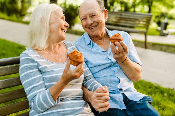 White senior couple smiling and eating muffins while sitting on bench in summer park