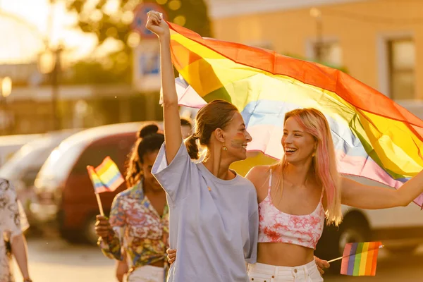 若いレズビアンカップル笑顔と歩くと虹の旗でプライドパレード中に街の通り — ストック写真