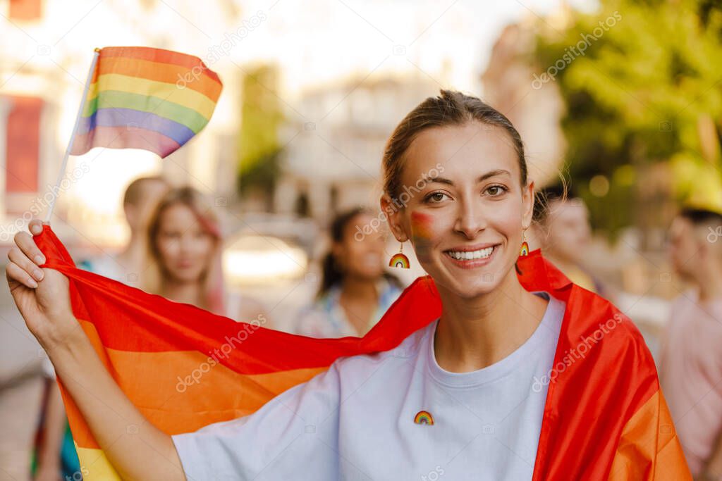 Young lesbian woman wrapped in rainbow flag smiling during pride parade on city street
