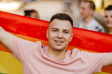 Elinde gökkuşağı bayrağı olan gülümseyen genç bir adama yakın durun. Bir lgbt topluluğunun geçit töreninde gülümseyen farklı insanların arasında ayakta durun.
