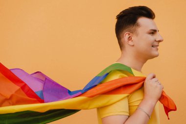 Sarı duvarın üzerinde duran, gülümseyen ve elinde gökkuşağı bayrağı tutan genç eşcinsel adam.