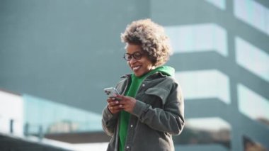 Günlük giysiler içinde gülen Afrikalı kadın. Açık havada telefonla mesaj atıyor.