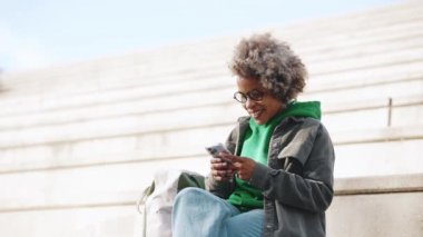 Pozitif yetişkin Afrikalı kadın. Günlük kıyafetler giyip telefonda yazıyor. Merdivenlerde oturuyor.