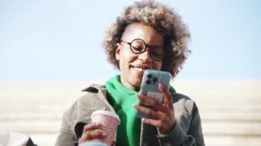 Neşeli, yetişkin Afrikalı kadın. Günlük kıyafetler giyip telefona bakıyor ve merdivenlerde kahve içiyor.