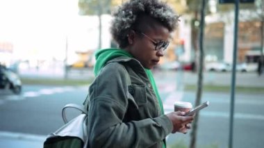 Ciddi bir Afrikalı kadın. Günlük giysiler içinde. Şehirde yürürken telefona bakıyor.