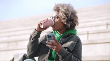 Sıradan kıyafetler giyen, telefona bakan ve merdivenlerde oturup kahve içen pozitif Afrikalı kadın.