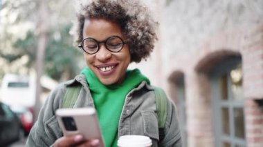 Yeşil kapüşonlu giyen neşeli Afrikalı kadın dışarıda telefonla mesajlaşıyor.