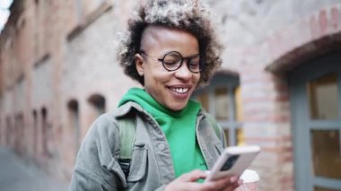 Yeşil kapüşonlu giyen gülümseyen Afrikalı kadın telefonla mesaj atıyor ve dışarıda yürüyor.