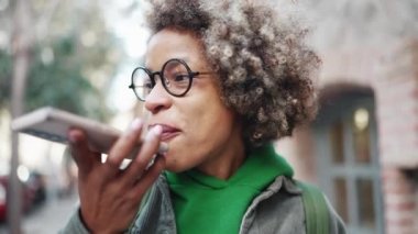 Yeşil kapüşonlu gülen Afrikalı kadın açık havada telefonla sesli mesaj kaydediyor.
