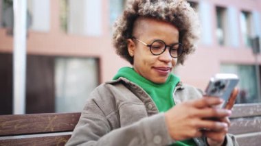 Düşünceli Afrikalı kadın yeşil kapüşonlu, akıllı telefonla yazı yazan ve dışarıdaki kameraya bakan.